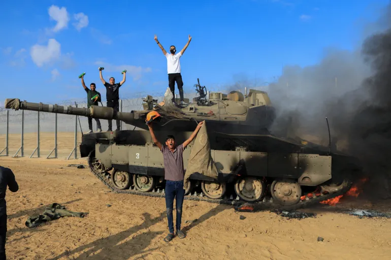 Palestinians celebrate on the Israeli side of the Israel-Gaza border where an Israeli tank burns after it was attacked by Palestinian fighters [Yasser Qudih/Reuters]